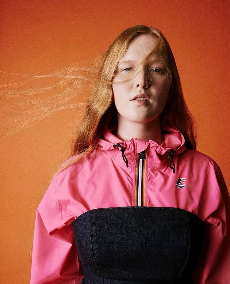 Miriam Rochholz in der K-Way-Kampagne: Porträt in pinker Regenjacke vor orangefarbenem Hintergrund, das rote Haar weht ins Gesicht