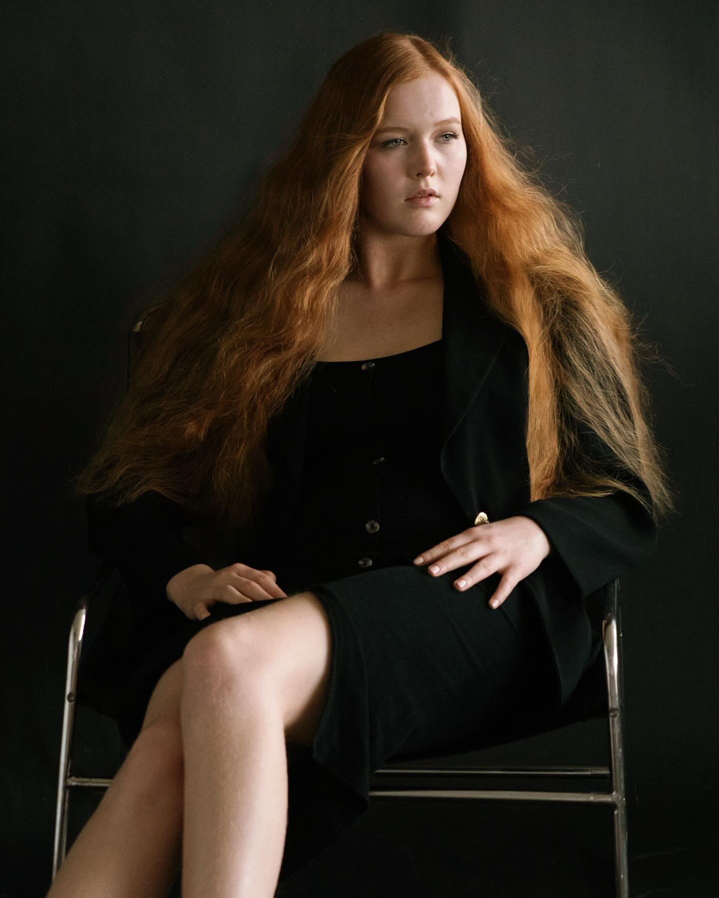 Miriam Rochholz sitzt in schwarzem Blazer und Kleid auf einem Stuhl vor schwarzem Hintergrund, das lange rote Haar offen