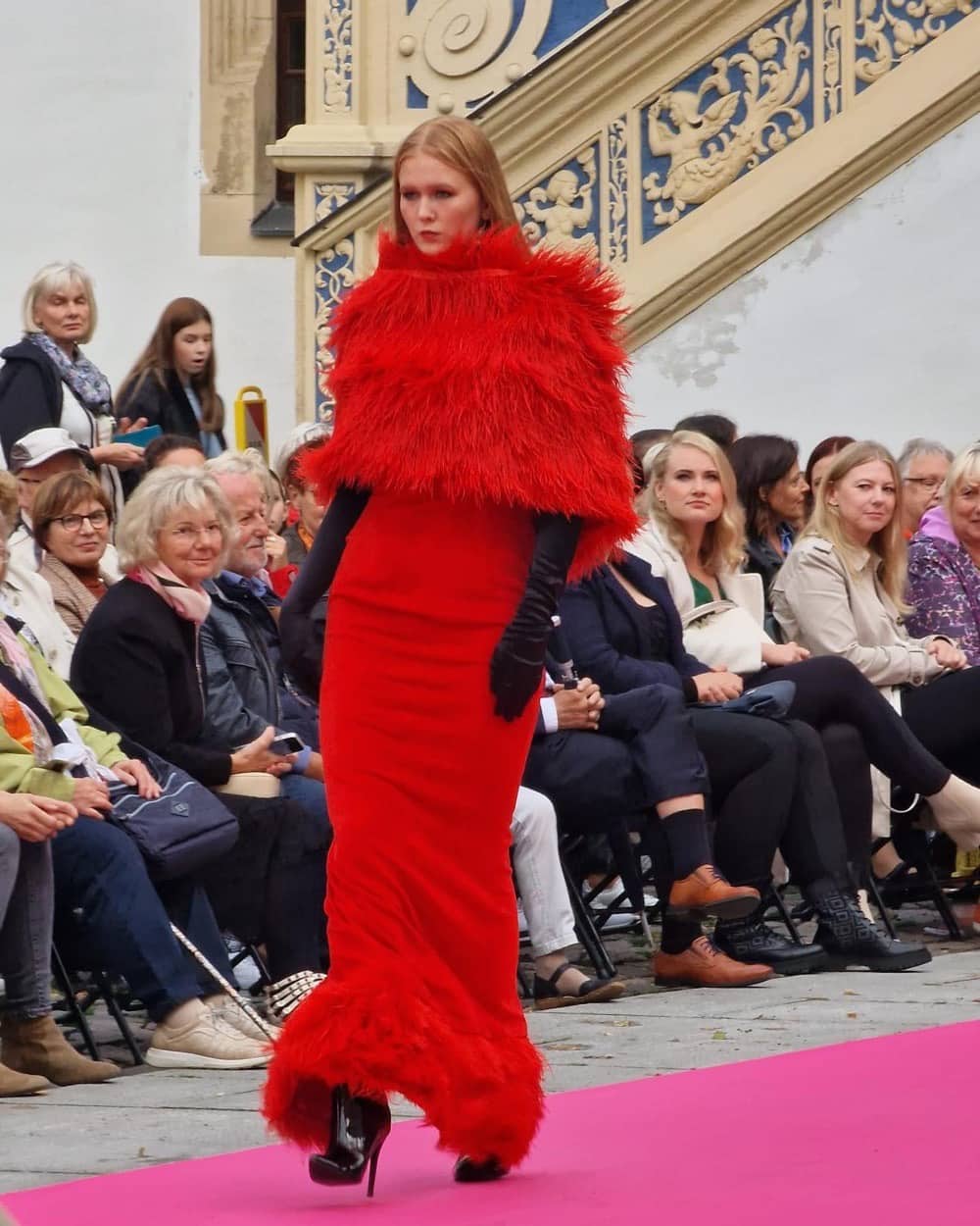 Miriam Rochholz auf einem pinkfarbenen Runway in einer roten Robe mit Federbesatz von Nardou Couture, Publikum am Rand